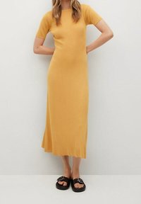 Robe tricotée jaune moutarde à manches courtes avec une silhouette ajustée, longueur mi-mollet, associée à des sandales noires avec un détail de boucle.