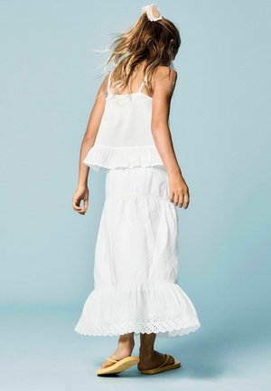 Hosszú hajú lány, aki fehér ujjatlan felsőt és fodros szoknyát visel, fodros szegéllyel, bézs szandálokat és virágos hajcsatot, világoskék padlón áll.