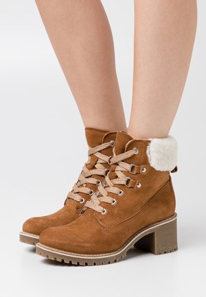 Botines de ante marrón que presentan un puño de piel sintética, ojales de metal y cordones beige, con un tacón grueso texturizado y suela de goma.