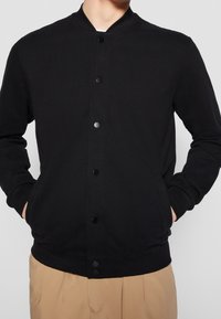 Veste bomber noire à boutons-pression en tissu doux, dotée d'un col, de poignets et d'un ourlet côtelés. Elle présente une texture simple et lisse, sans motifs.