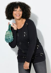 Zwarte top met lange mouwen voorzien van paillettenaccenten, gecombineerd met een groene handtas met franjes, gedragen met donkergrijze spijkerbroek.
