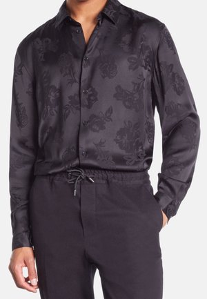 Męska czarna satynowa koszula na guziki z subtelnym kwiatowym wzorem, włożona do czarnych spodni na sznurku, jedna ręka w kieszeni.