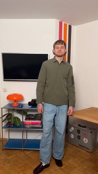 Muž v zelené bundě a modrých džínách stojí v místnosti s televizí, oranžovou lampou, fotoaparátem, knihami a kufrem označeným Tropicana.