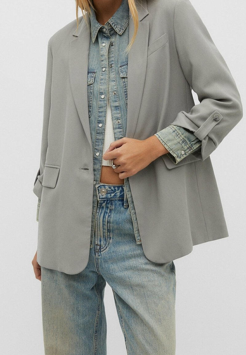 Femme portant un blazer gris clair par-dessus une chemise en jean et un jean, avec la main ajustant le blazer près de la taille.