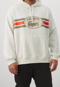 Felpa bianca con tasca a scomparsa, con un grande logo Lacoste e strisce orizzontali in marrone e rosso sul petto.