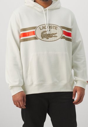 Felpa bianca con tasca a scomparsa, con un grande logo Lacoste e strisce orizzontali in marrone e rosso sul petto.
