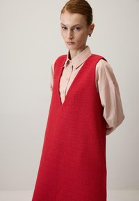 Červené textúrované bezrukávové šaty s výstrihom do V, navrstvené na svetloružovej košeli s golierikom. Majú voľný strih a minimalistický dizajn.