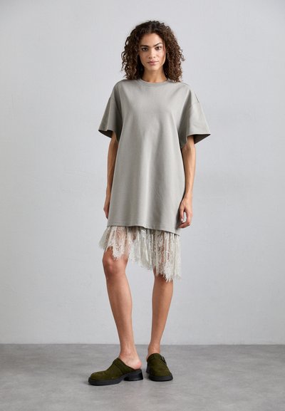 Robe t-shirt grise surdimensionnée avec des manches courtes et une garniture en dentelle à l'ourlet. Le modèle porte des sabots vert foncé. L'arrière-plan est un mur gris clair.