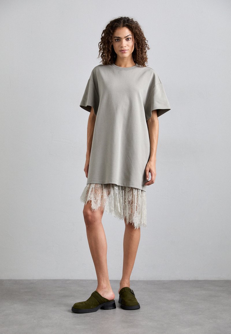 Hallikas üle mõõtme t-särk-kleit lühikeste varrukatega ja pitsiga ääres. Mudel kannab tumerohelisi klotse. Taust on helehall sein.