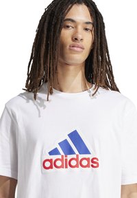 T-shirt bianco in cotone con una grande grafica del logo Adidas blu e rosso sul davanti. Collo a giroclassico e maniche corte.