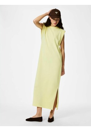 Mujer con vestido midi sin mangas de color amarillo pálido con abertura lateral, gorra marrón y zapatos planos negros, posando con una mano en la cabeza.