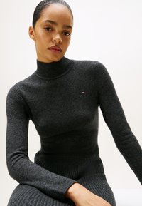 Sivi turtleneck pulover izrađen od mekog pletenog materijala s uskim krojem. Ima duge rukave i mali logo detalj na prsima.