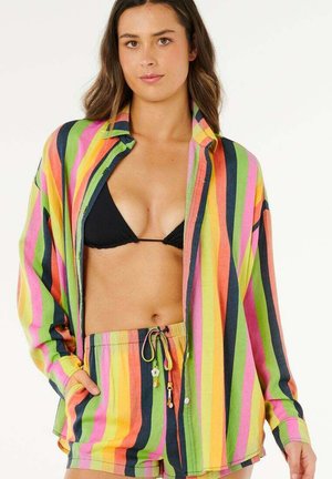 Mujer con una camisa de rayas verticales coloridas y pantalones cortos a juego con un bikini negro debajo, de pie con una mano en el bolsillo.