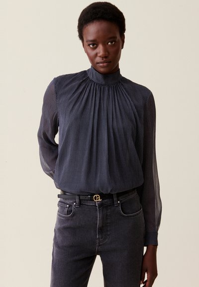 Blusa de color navy con pequeños lunares blancos, cuello alto con pliegues y mangas largas transparentes. Combinada con jeans oscuros de mezclilla y un cinturón negro.