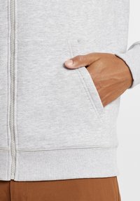 Sweatshirt zippé gris en tissu texturé, doté d'une poche avant et de poignets et d'un ourlet côtelés, associé à un pantalon marron.