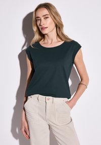 Tmavě zelené tričko s krátkým rukávem a kulatým výstřihem, spárované se světle béžovými kalhotami s vysokým pasem a předními kapsami.