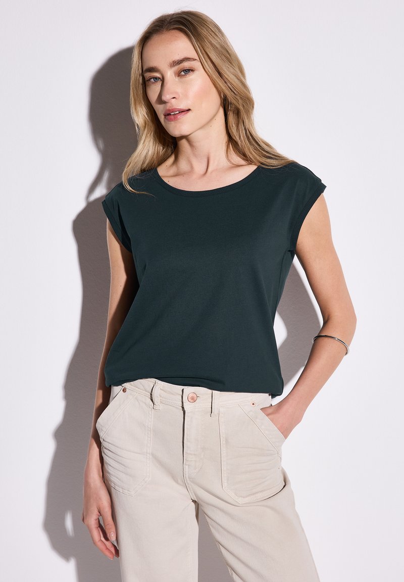 Tmavě zelené tričko s krátkým rukávem a kulatým výstřihem, spárované se světle béžovými kalhotami s vysokým pasem a předními kapsami.