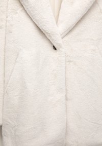 Zbliżenie miękkiego, kremowego futra sztucznego z szerokimi klapami i pojedynczym widocznym guzikiem przy kołnierzu.