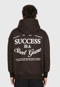 Personne portant un sweat à capuche noir avec un texte blanc au dos : "Enseigné par les rues. Le succès est un jeu de rue. Rêvez grand, travaillez dur et réalisez-le."