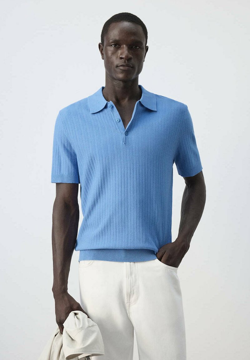 Homme portant une chemise en maille bleu clair à manches courtes avec col et un pantalon blanc, tenant une veste légère pliée dans une main.