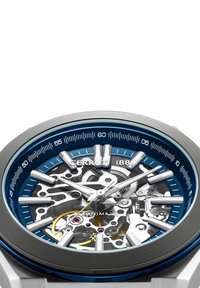 Skeleto laikrodis su mėlynu rėmeliu, sidabriniais akcentais ir skaidriu ciferblatu, atskleidžiančiu mechaninius komponentus ir auksinį ratelį. Dideli žymekliai, kad būtų lengviau perskaityti.