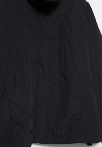 Musta kevyt takki, jossa on korkea kaulus ja näkyvät sauma- ja ompelualueiden yksityiskohdat yksivärisellä taustalla.