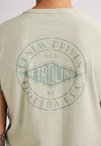 Světle zelené tričko s krátkým rukávem, které má velký kruhový grafický prvek s textem "DENIM DRIVEN" a "PETROLIND" v retro stylu.