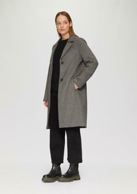 Manteau en laine gris avec col cranté, deux boutons noirs, poches latérales, associé à un pull noir texturé et un pantalon palazzo noir. Bottes noires.