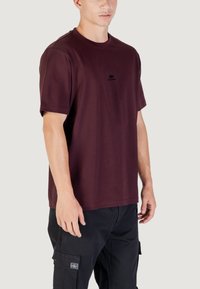 Camiseta corta de manga en color burdeos, hecha de tela suave, con un logo negro en el pecho. Combinada con pantalones cargo negros con bolsillos.