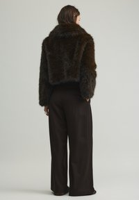 Giacca corta in pelliccia sintetica marrone con colletto ampio, abbinata a pantaloni neri a gamba larga e vita alta, con superficie testurizzata.