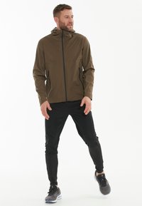 Veste légère vert olive avec capuche, fermeture éclair à l'avant et accents noirs ; associée à un pantalon noir ajusté et des chaussures de sport.