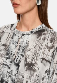 Durchsichtige Bluse mit detailliertem Schwarz-Weiß-Stadtbilddruck, Rüschenausschnitt und Knopfverschluss vorne. Texturierter Stoff.