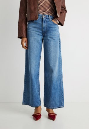 Persoană îmbrăcată cu blugi albaștri cu talie înaltă și picioare largi, geacă din piele maro, cămașă în carouri și pantofi roșii cu vârf ascuțit, stând pe un fundal simplu.