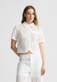 Camicia bianca a maniche corte con orlo scallop, caratterizzata da dettagli in pizzo ad occhio, colletto a punta e tasca frontale. Abbinata a pantaloni coordinati.