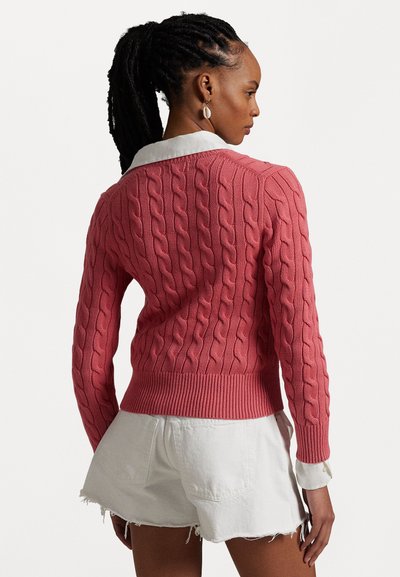 Rosa Kabelstrickpullover mit geripptem Saum und Kragen über weißen Hemdfällen, kombiniert mit weißen Jeansshorts.