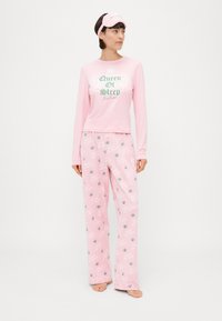 Conjunto de pijama rosa com uma blusa de mangas longas apresentando a inscrição "Rainha do Sono" e calças correspondentes com um padrão repetido de coroas e texto.