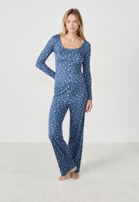 Kobieta stoi boso w niebieskiej, kwiatowej bluzce z długim rękawem zapinanej na guziki oraz dopasowanych szerokich spodniach, na tle białego tła.