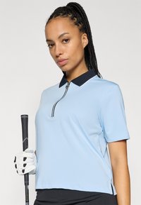 Svijetloplava polo majica s tamnoplavim ovratnikom, kratkim rukavima i zaključavanjem na četvrtinu. Ruka prekrivena rukavicom drži crni rukohvat golf palice.