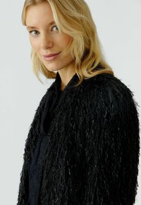Blonde Frau mit hellen Augen, die eine strukturierte schwarze, fellartige Jacke trägt. Sie schaut leicht zur Seite und hat ein dezentes Lächeln auf den Lippen, vor einem schlichten Hintergrund.