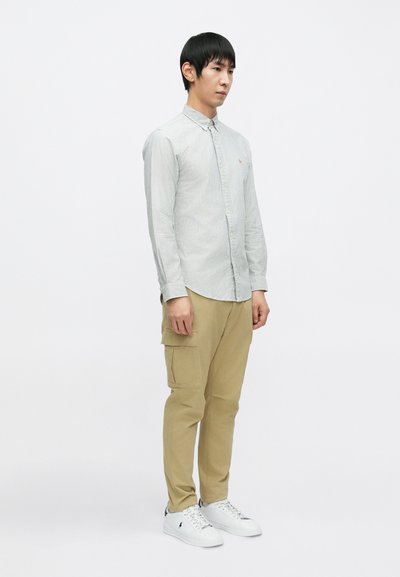 Pruhovaná košile na knoflíky světle šedé barvy, béžové kalhoty s kapsami a bílé tenisky. Košile má na prsou malou logo.