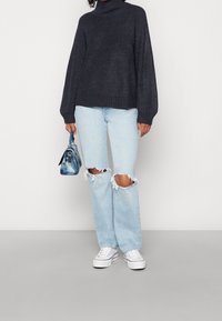 Mörkgrå stickad turtleneck-tröja, looser passform, kombinerad med ljusblå rivna jeans och vita sneakers. Håller en blå tie-dye-handväska.