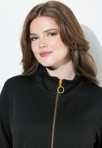 Chaquetón negro con un sutil patrón animal, cuello alto y cremallera amarilla con un tirador circular. Presenta una tela texturizada.