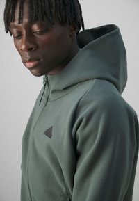 Sweatshirt à capuche verte avec fermeture éclair entièrement, fabriqué à partir d'un matériau doux. Présente un petit logo noir sur la poitrine, sans motifs ni accents visibles.