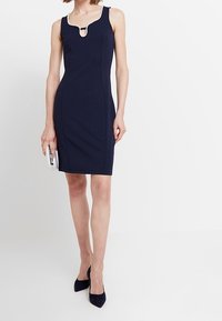 Granatowa dopasowana sukienka bez rękawów, z dekoltem w kształcie klucza ozdobionym perłowymi akcentami. Materiał wydaje się gładki i dobrze skrojony.