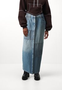 Jeans maxi szoknya foltos dizájnnal, világoskék és sötétkék árnyalatokkal, egyenes szabással és dísztűzéssel; fekete csizmával viselve.