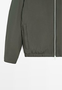 Giacca bomber verde oliva con chiusura frontale con cerniera, vita elastica, tasche laterali e tessuto liscio e opaco.