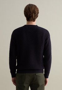 Marineblauwe gebreide trui met een gestructureerd patroon, geribde kraag en manchetten, en een relaxte pasvorm. Gemaakt in combinatie met olijfgroene broek.