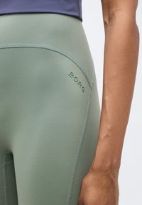 Κοντινό πλάνο ενός ατόμου που φορεί ανοιχτό πράσινο κολάν με το λογότυπο «BORG» κοντά στο ισχίο και εμφανή ραφή, με το χέρι χαλαρά στο πλευρό.