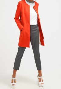 Červený šitý kabát s hladkou texturou, vrstvený přes bílý top a šedé kalhoty. Obsahuje bílé platformové sandály se strappy.