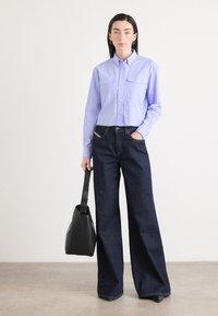 Camicia colletto viola chiaro con dettagli in bottone, abbinata a jeans a gamba larga blu scuro. Il modello tiene in mano una borsa in pelle nera.
